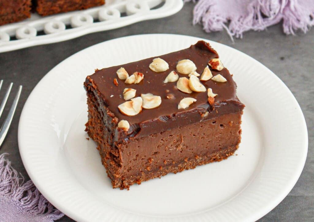 A slice of chocolate hazelnut cake topped with chopped hazelnuts is served on a white plate.