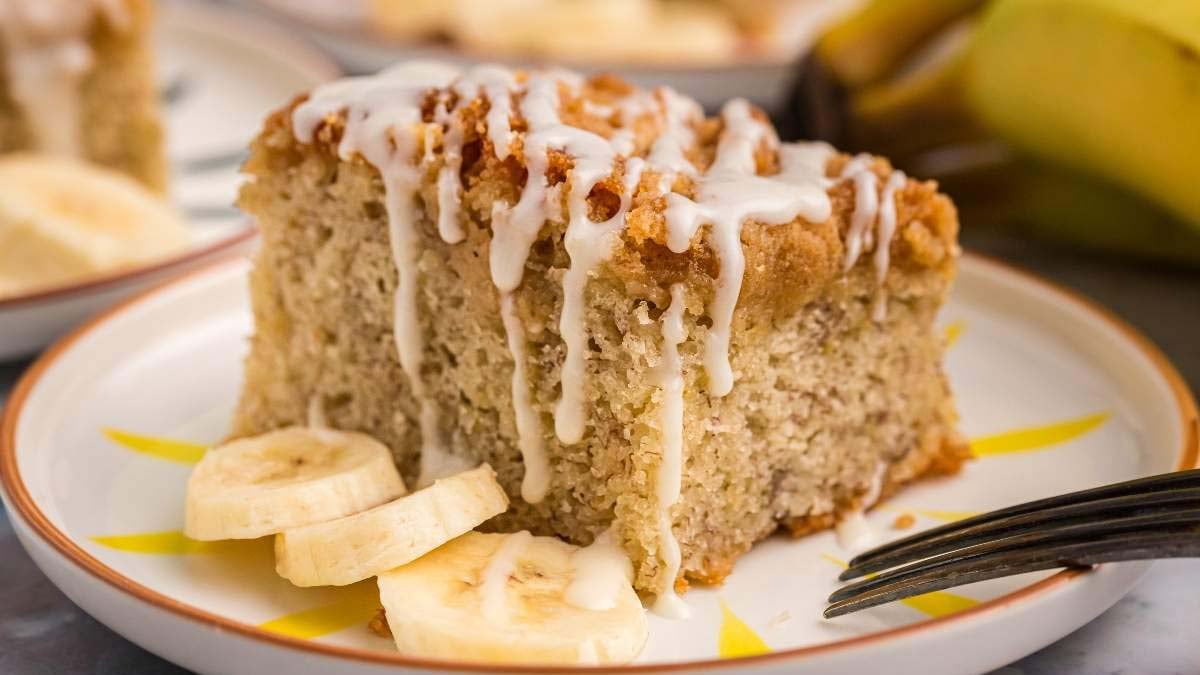 A slice of banana cake with white icing drizzled on top, served on a plate with banana slices and a fork.