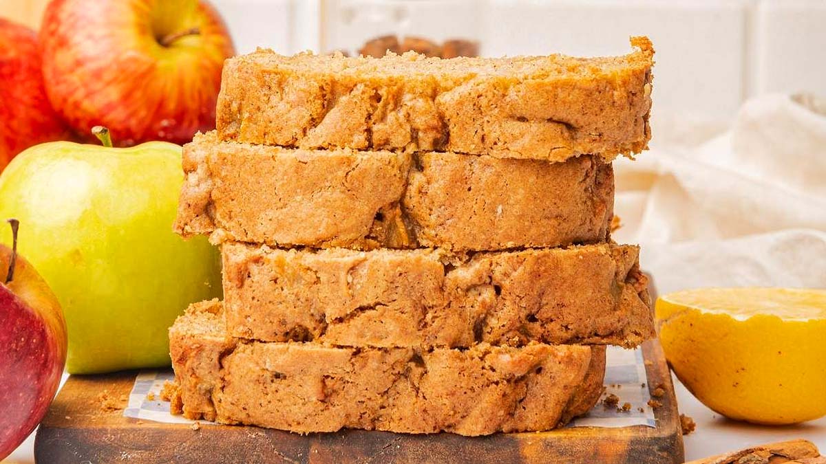 Four thick slices of apple bread are stacked on a wooden board, surrounded by whole apples and a half lemon in the background.