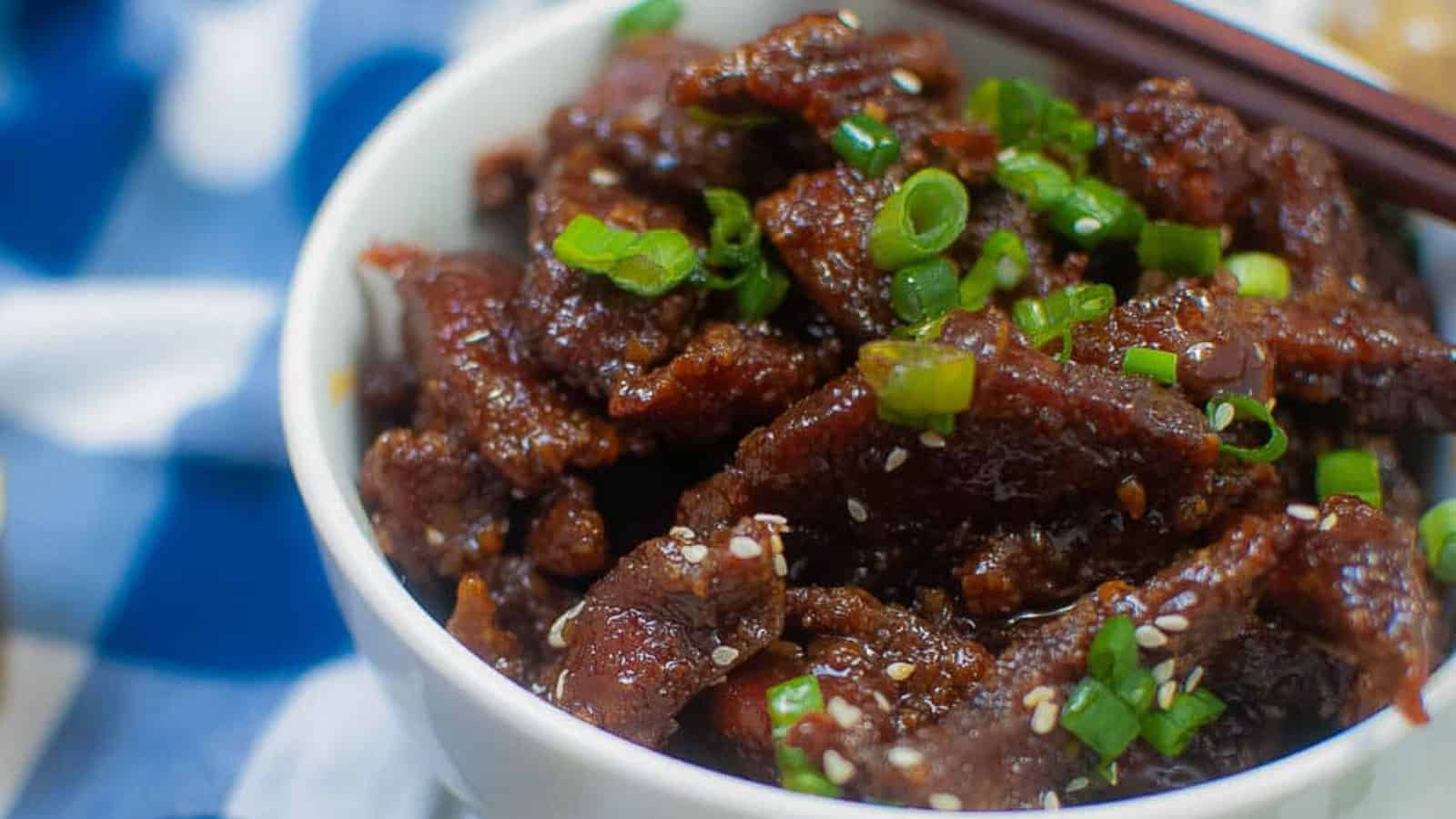 A white bowl filled with pieces of glazed beef topped with sesame seeds and chopped green onions.