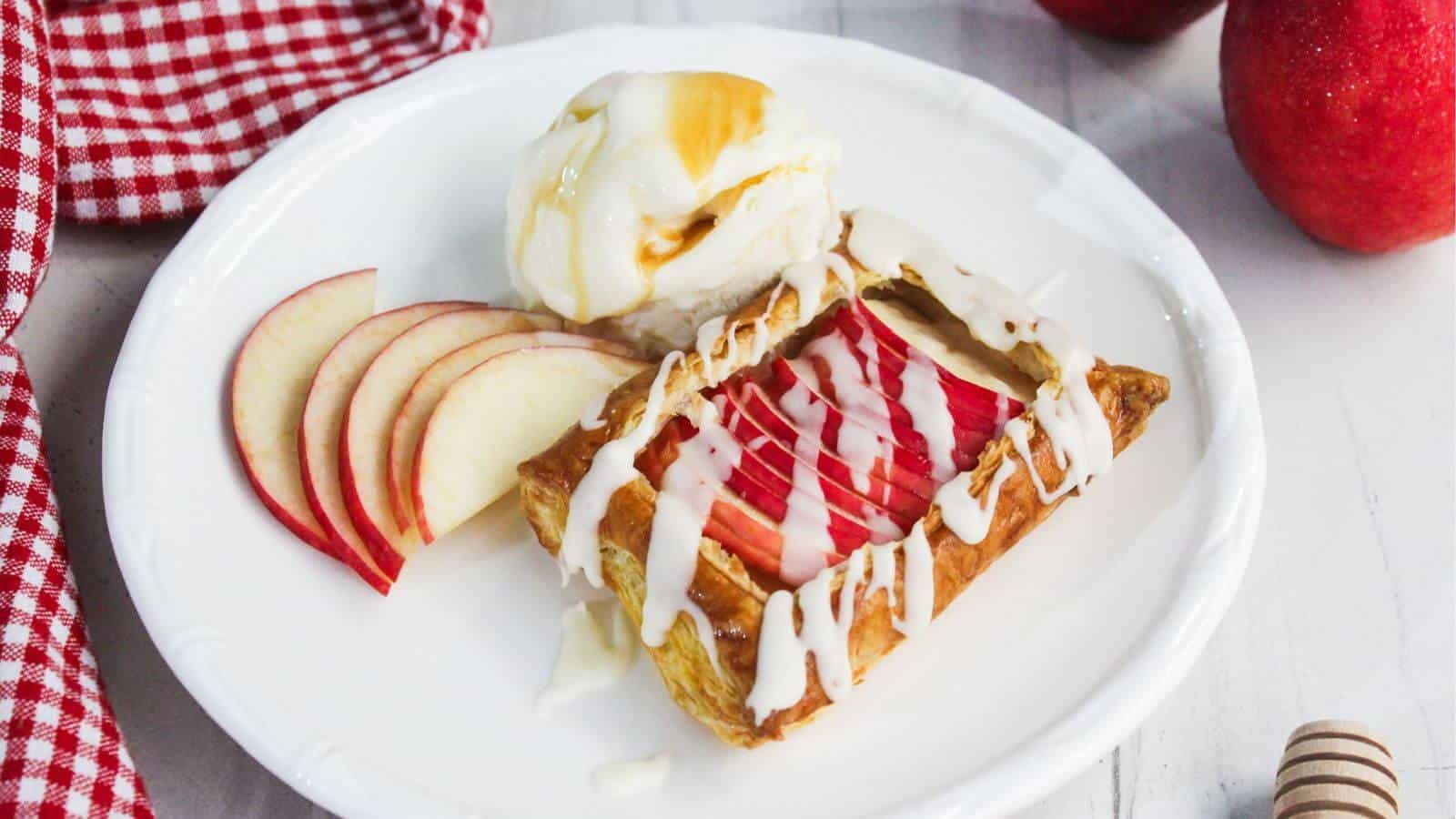 A plated dessert with a rectangular pastry topped with sliced apples and white icing, served with apple slices and a scoop of vanilla ice cream drizzled with syrup.