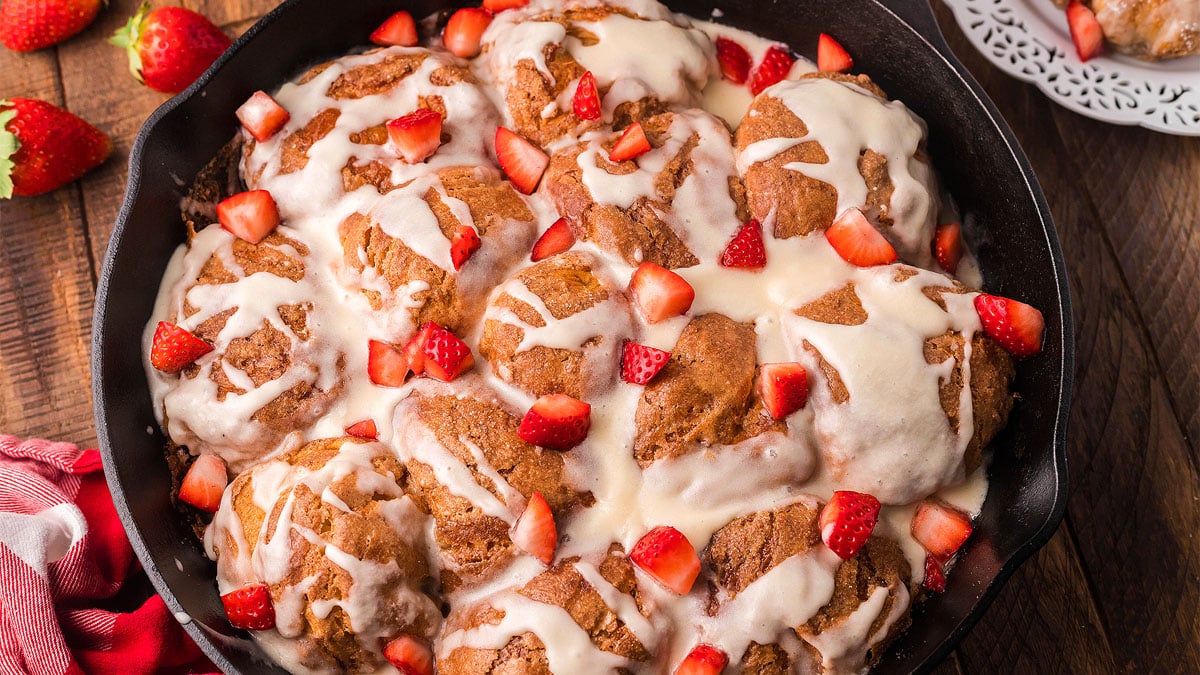 A skillet filled with baked cinnamon rolls topped with white icing and fresh chopped strawberries on a wooden table.