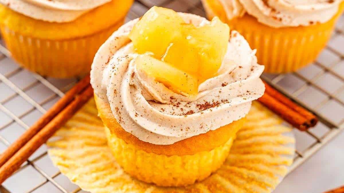 A cupcake topped with swirled frosting, cinnamon, and pineapple chunks, displayed on a wire rack with cinnamon sticks nearby.