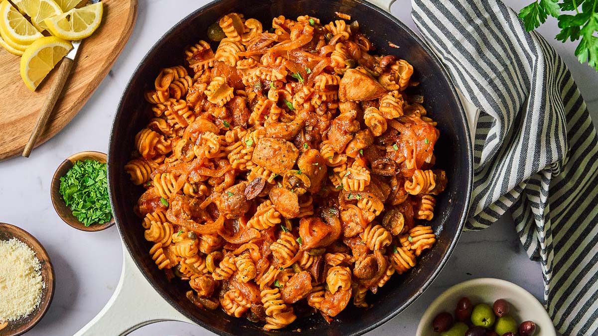 A skillet filled with rotini pasta, chicken, and tomato sauce sits on a table surrounded by lemon slices, grated cheese, chopped herbs, and olives.