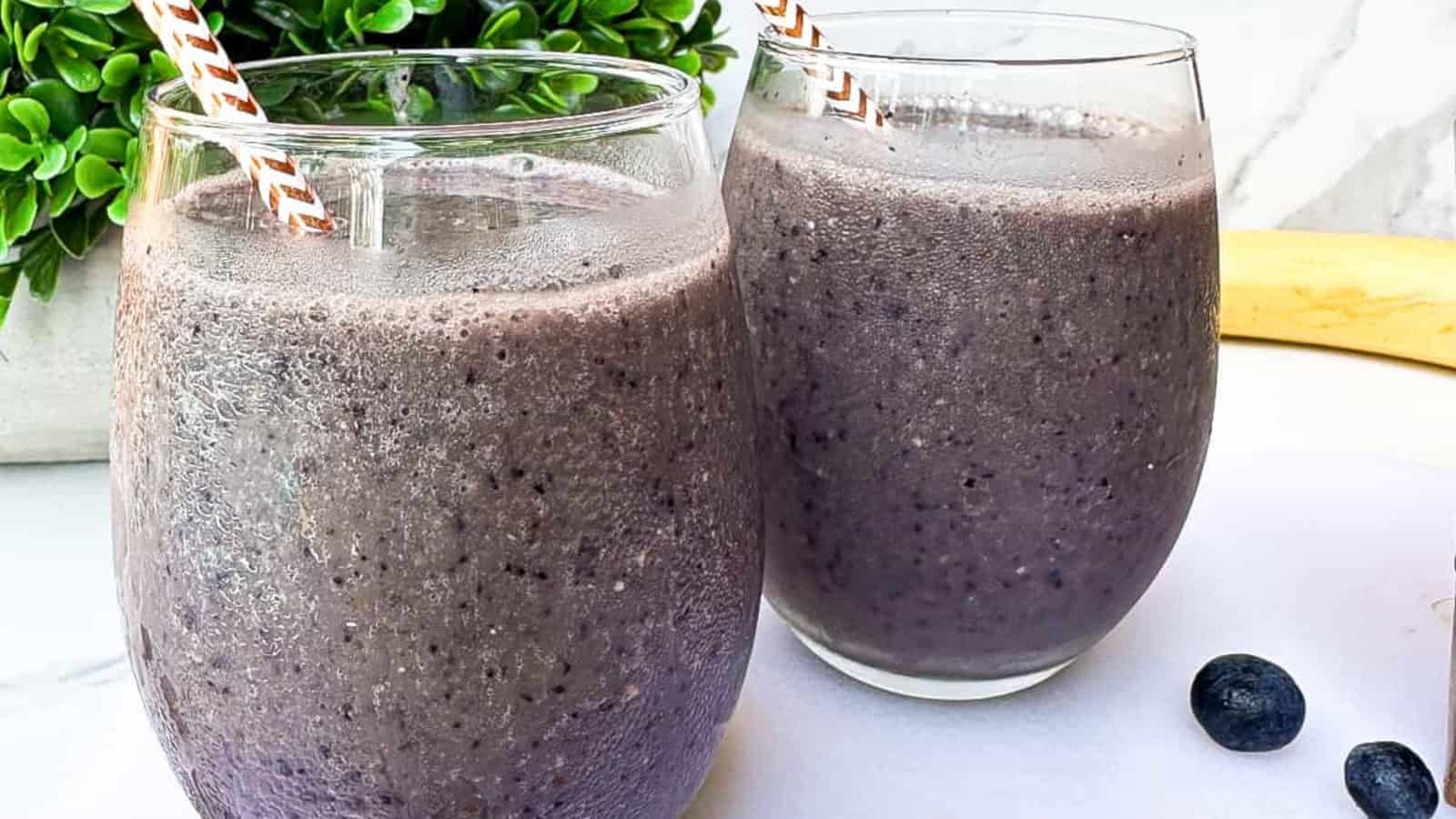 Two glasses of thick purple smoothie with straws, topped with condensation, placed on a white surface next to a few blueberries and a green plant.
