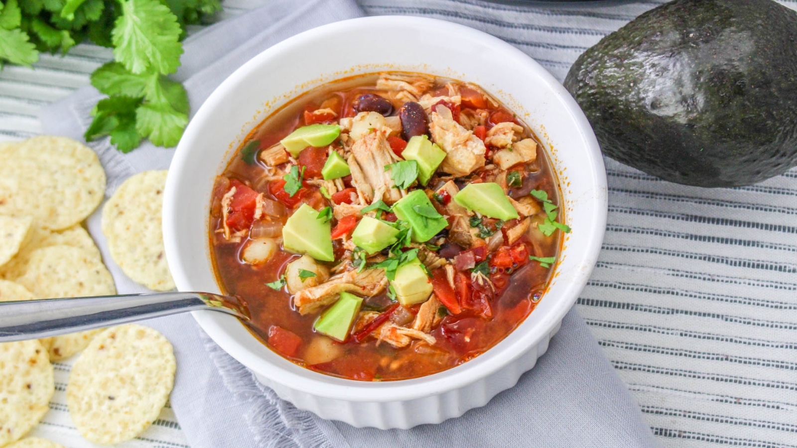 A bowl of chicken tortilla soup topped with diced avocado, with a spoon, tortilla chips, cilantro, and an avocado nearby on a striped cloth.