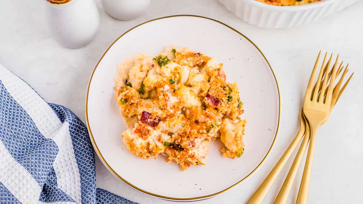 A serving of baked casserole with vegetables and breadcrumbs on a white plate, placed next to gold forks and a blue and white towel.