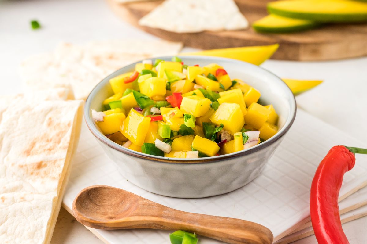 A bowl of mango salsa with diced mango, red onion, and peppers, next to a wooden spoon, tortilla pieces, and a red chili pepper on a white surface.