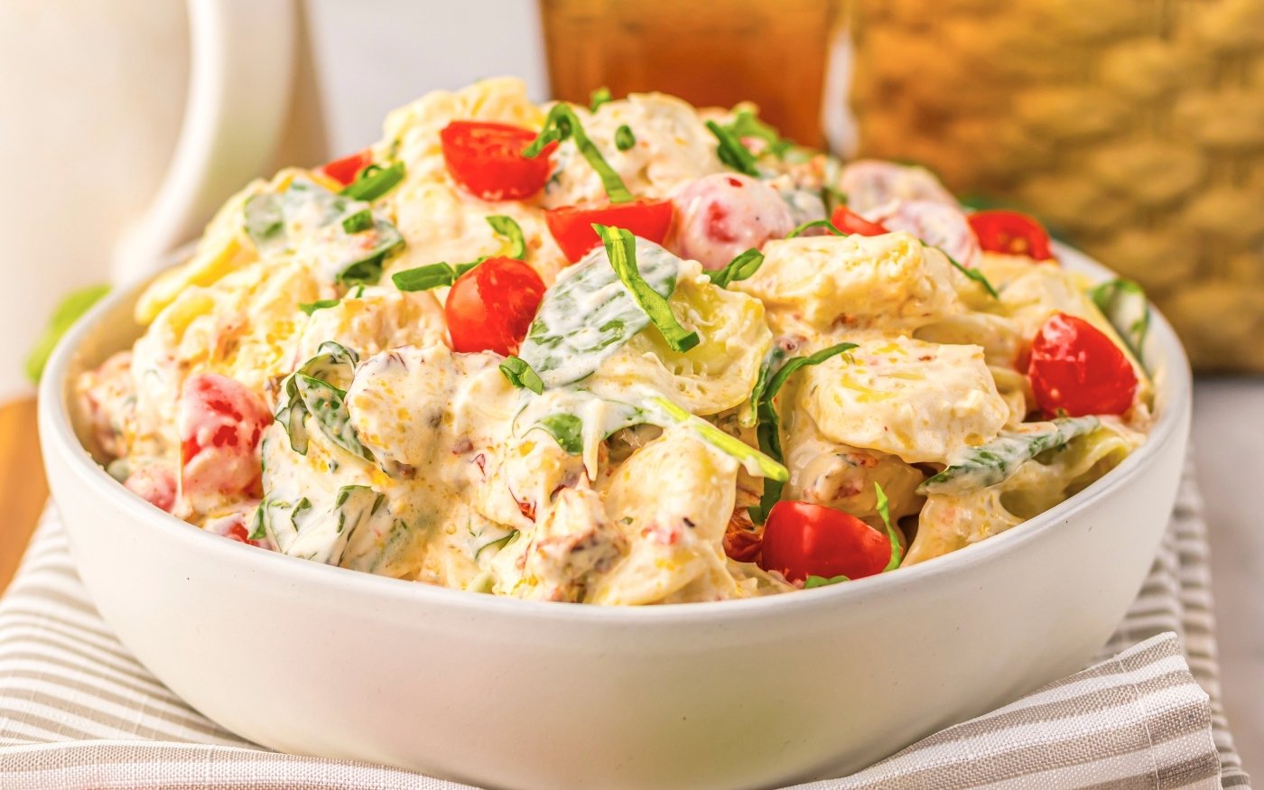 A white bowl filled with creamy potato salad, garnished with cherry tomato halves and chopped green onions, sits on a striped cloth.