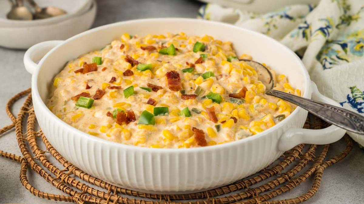 A white ceramic dish filled with creamy corn casserole topped with pieces of bacon and chopped green peppers, with a serving spoon on the side.