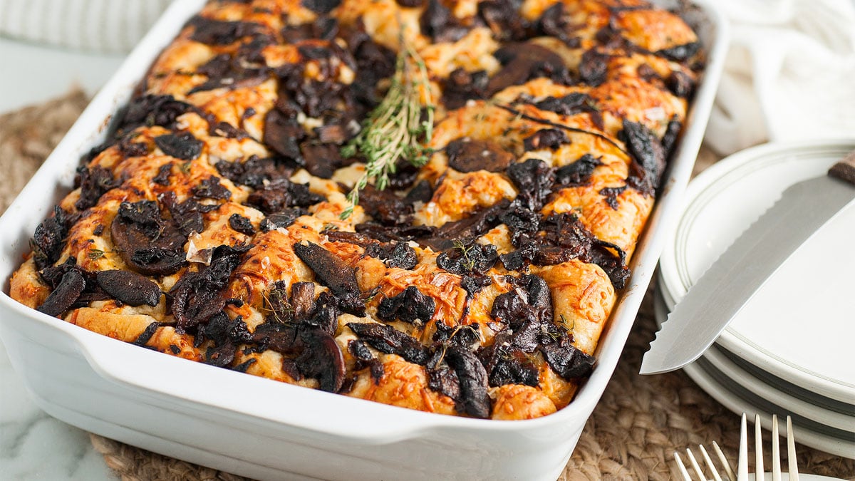 A baked casserole topped with cooked mushrooms and herbs in a white rectangular dish, placed next to plates, a knife, and a stack of forks.