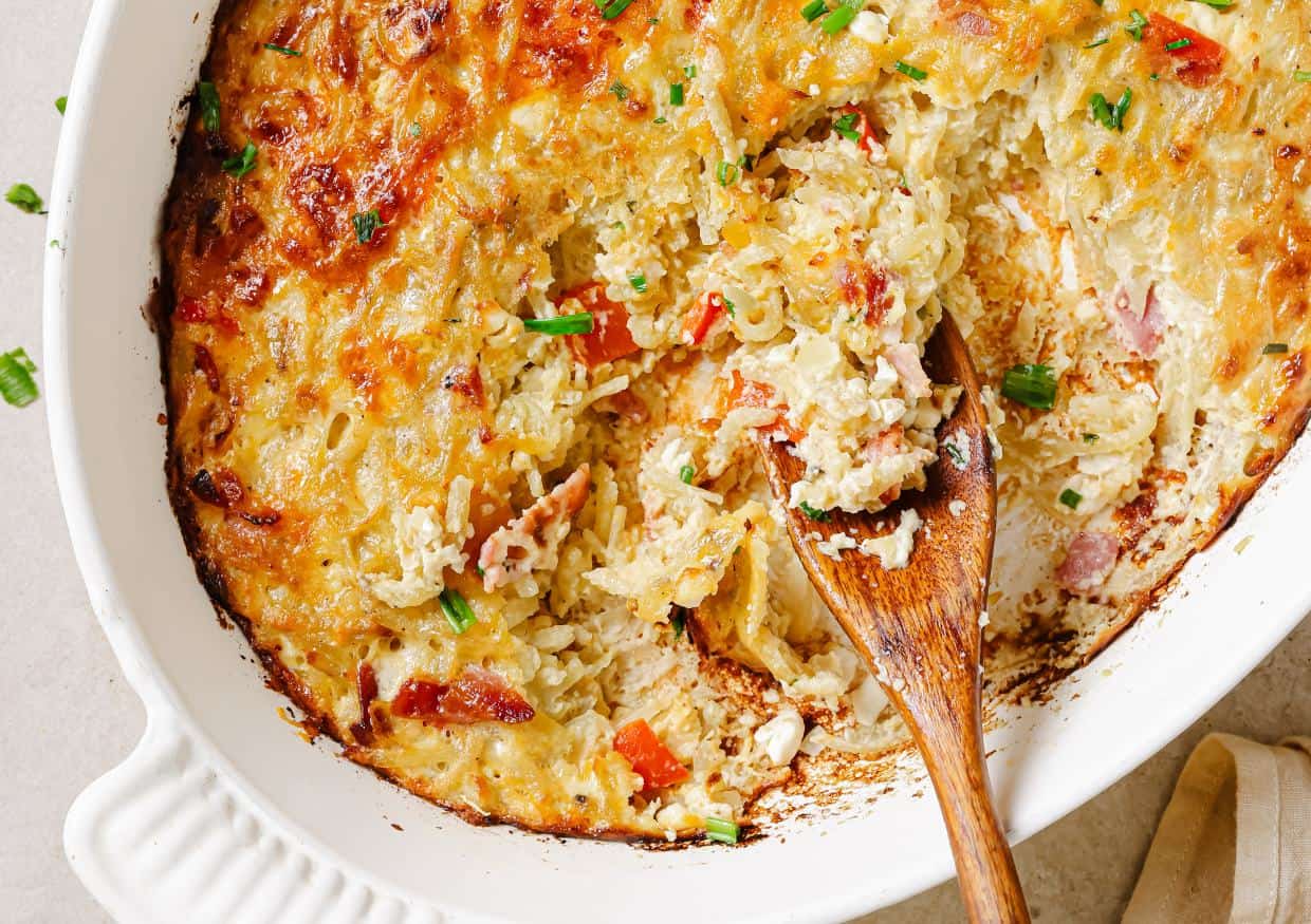 A baked casserole with a golden cheese crust, bits of bacon, and chives, partially served with a wooden spoon in a white dish.