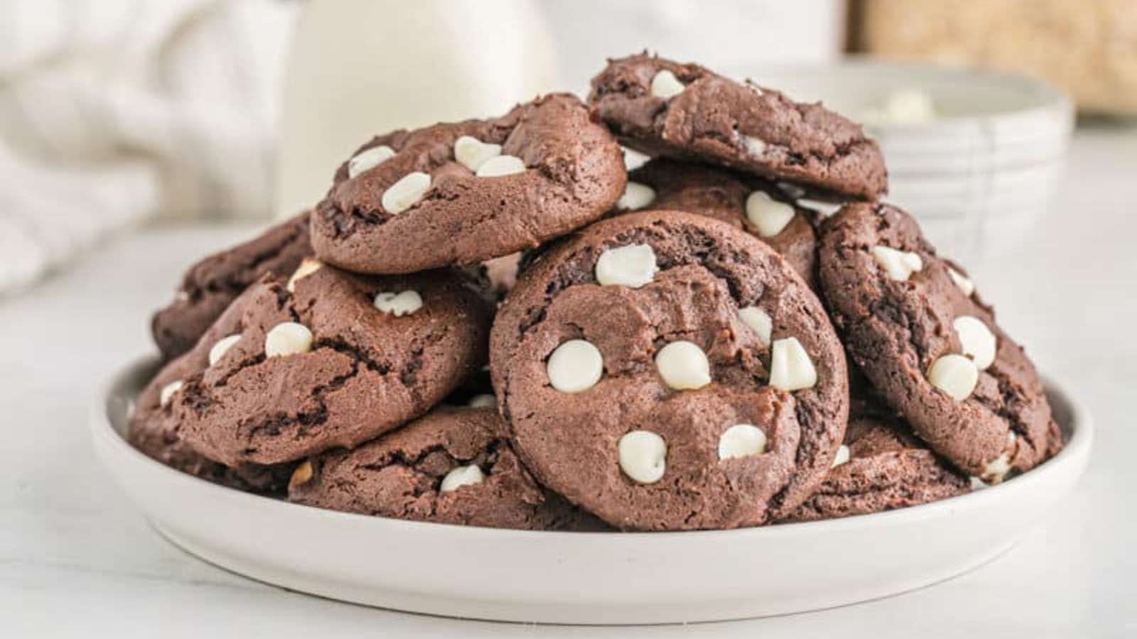 A white plate filled with stacked chocolate cookies with white chocolate chips on a light-colored surface.