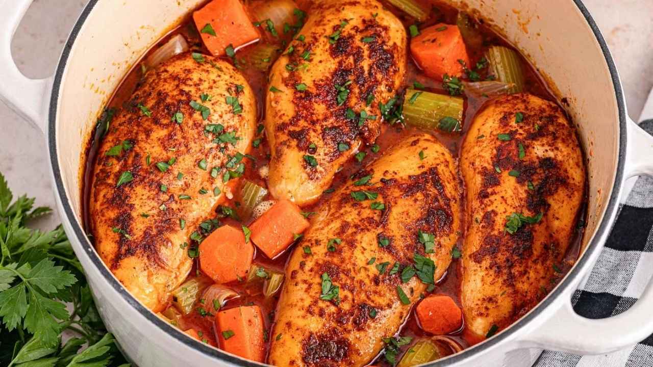 Four seasoned chicken breasts cooked with carrots and celery in a tomato-based broth inside a white Dutch oven.