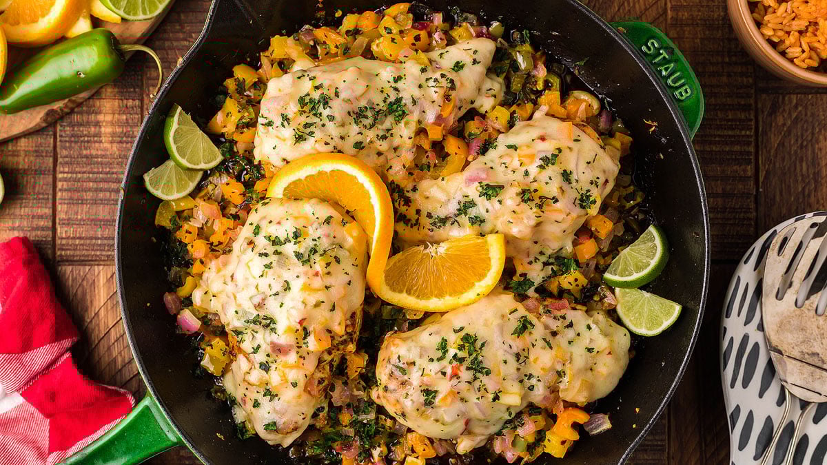 A skillet with cooked chicken breasts topped with herbs and orange slices, surrounded by chopped vegetables and lime wedges.