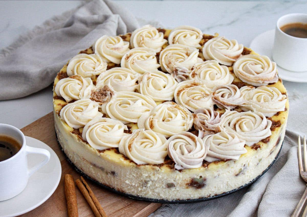 A round cheesecake topped with swirls of white and light brown frosting sits on a wooden board, next to two cups of coffee and a gray napkin.