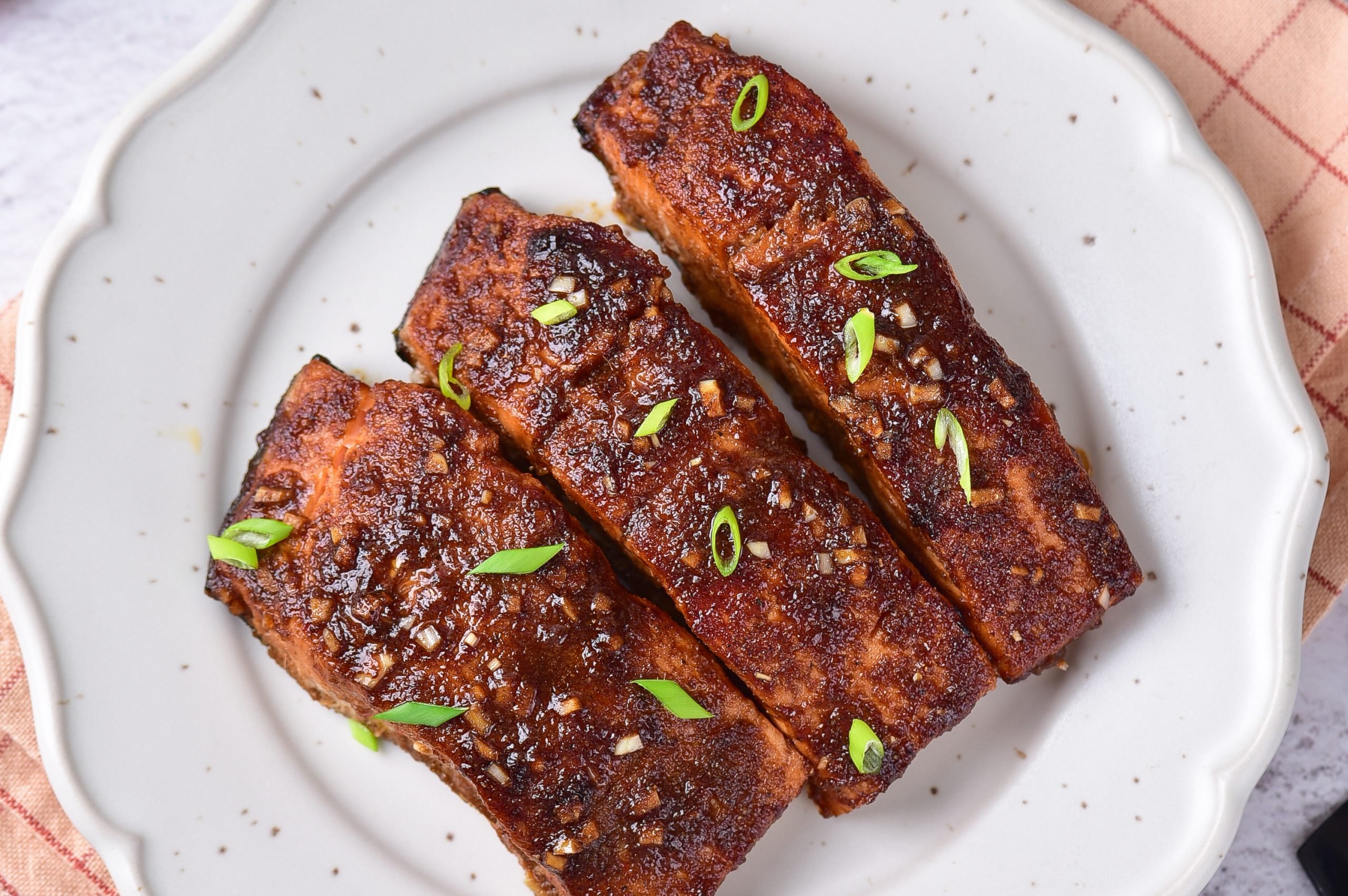 Three pieces of glazed salmon garnished with chopped green onions are arranged on a white plate.