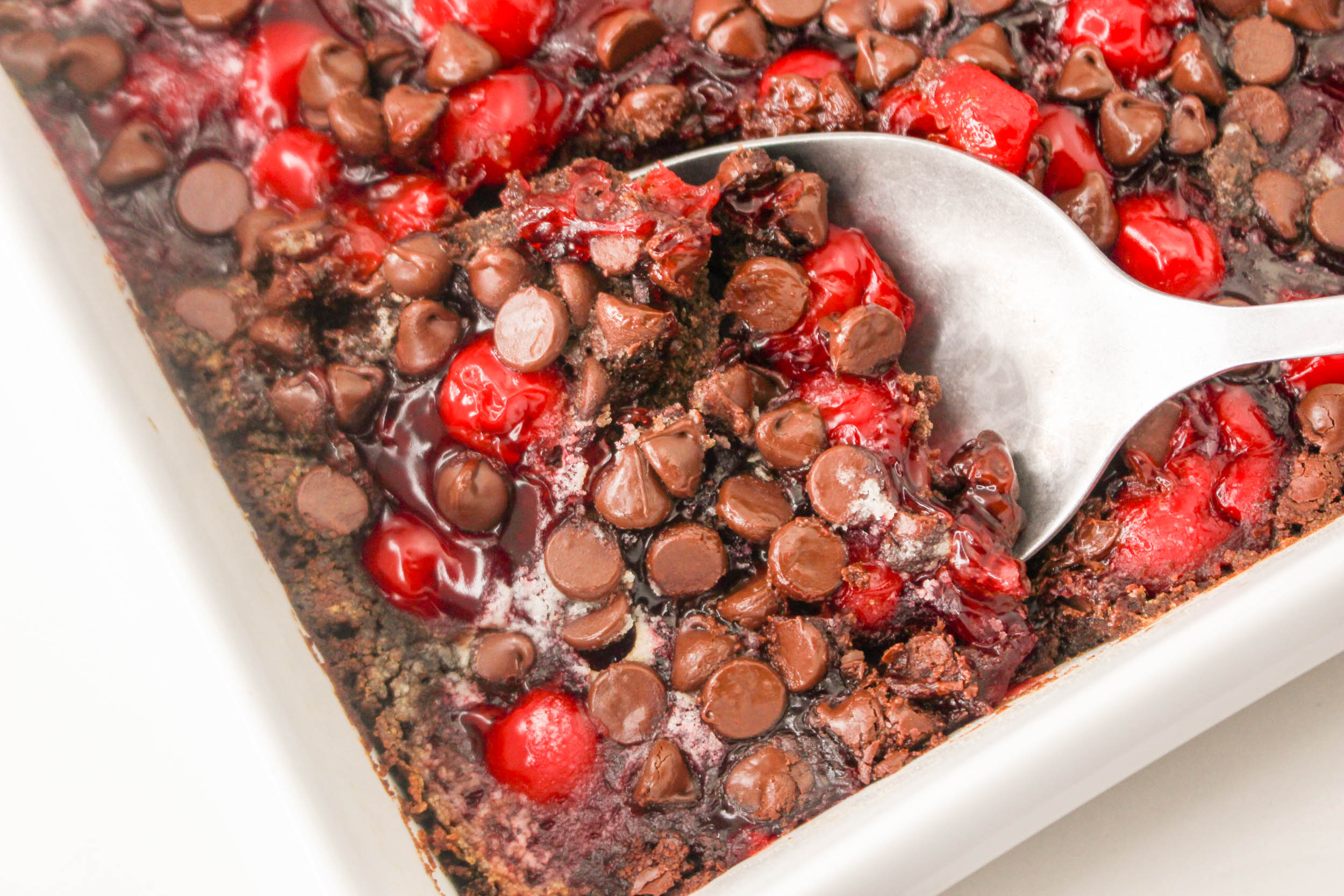 A baking dish with a chocolate cherry dessert topped with chocolate chips, with a serving spoon lifting a portion from the corner.