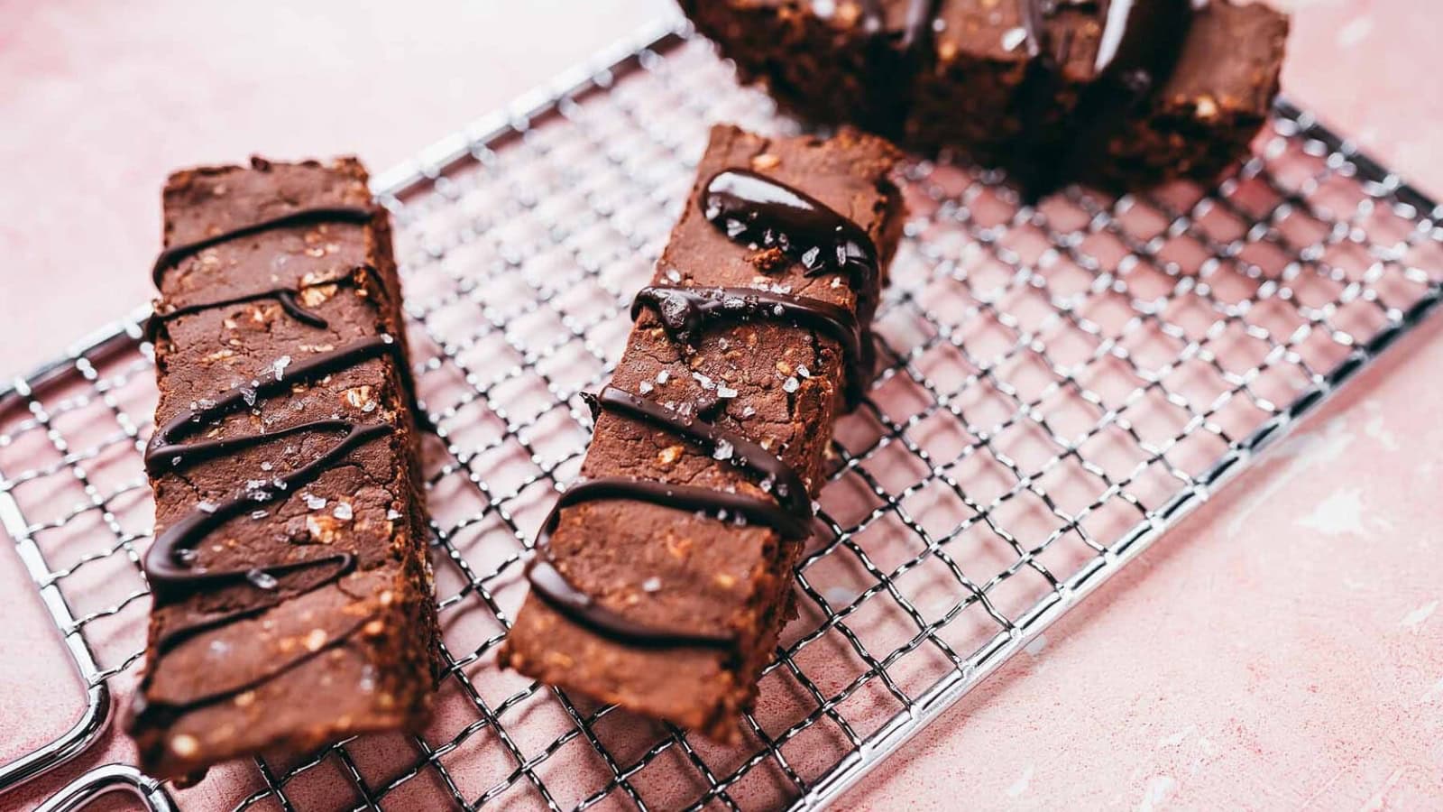 Two chocolate protein bars drizzled with chocolate sauce and sprinkled with sea salt, resting on a metal cooling rack.