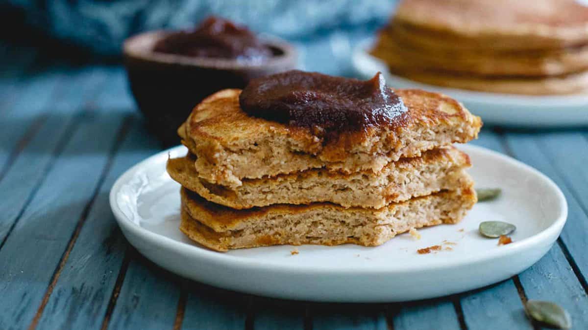A stack of three pancakes topped with a dollop of dark fruit spread on a white plate, with more pancakes and spread in the background.