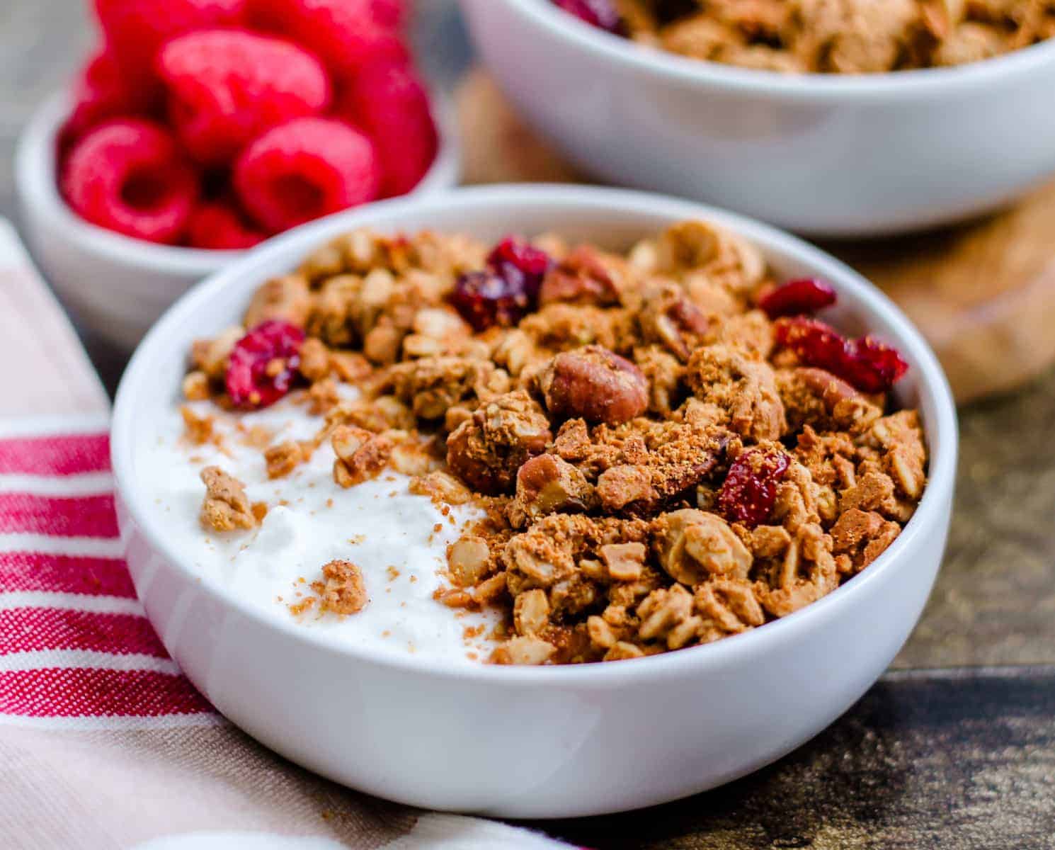 A bowl of yogurt topped with granola and dried fruit sits on a table next to a dish of fresh raspberries.