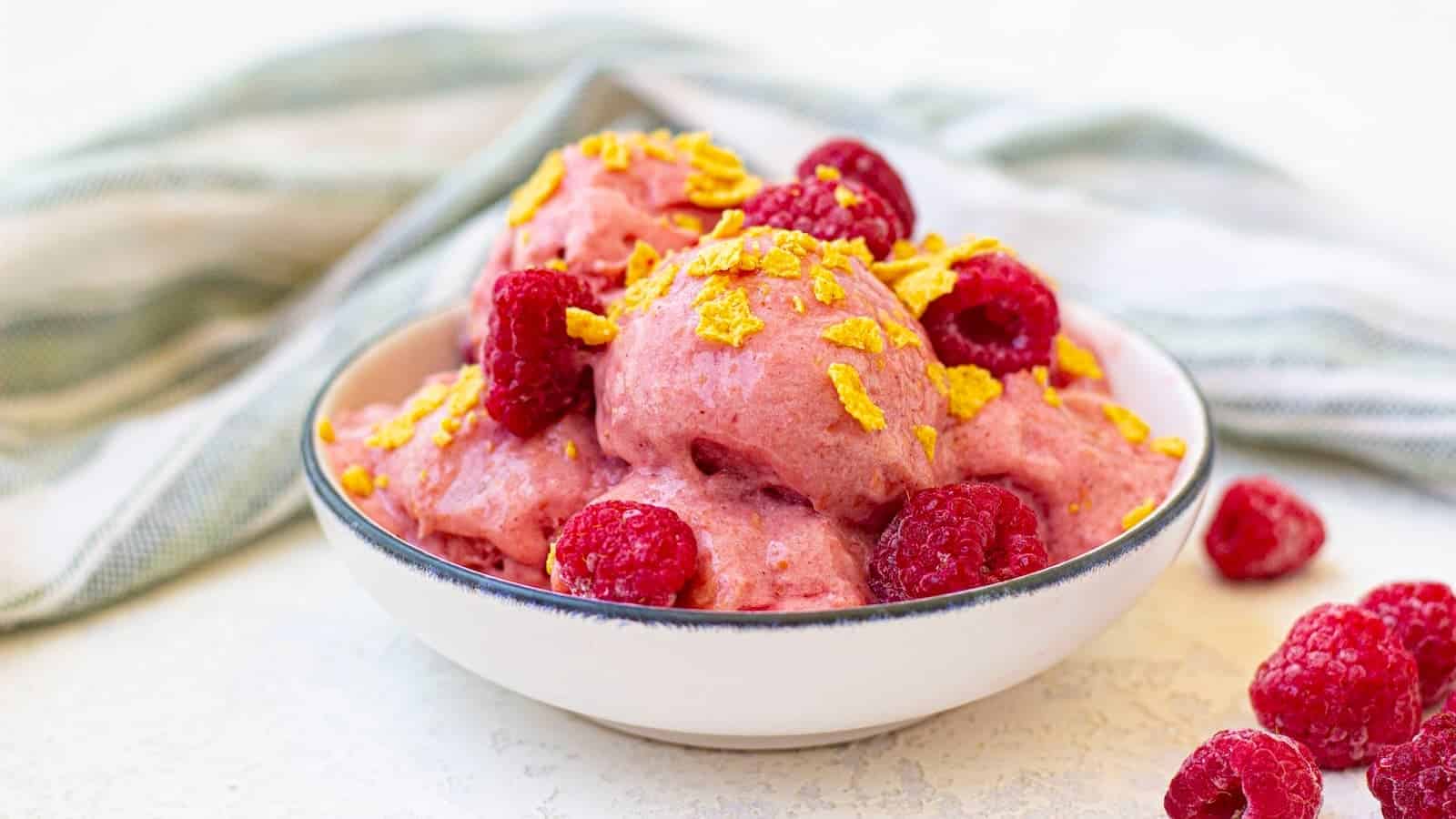 A bowl of pink raspberry ice cream topped with fresh raspberries and cornflake pieces, placed on a light surface with a striped cloth in the background.