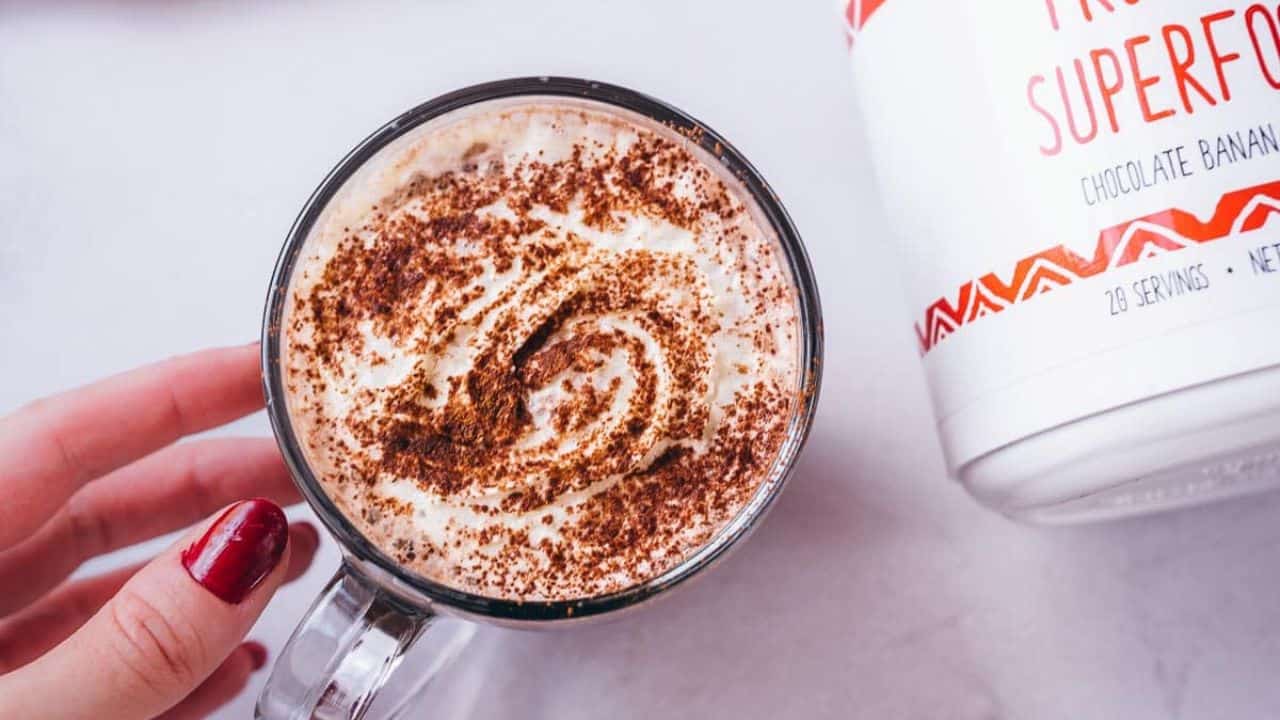 A hand holds a glass mug filled with a frothy beverage topped with cocoa powder; a container labeled “Superfood” is partially visible on the right.