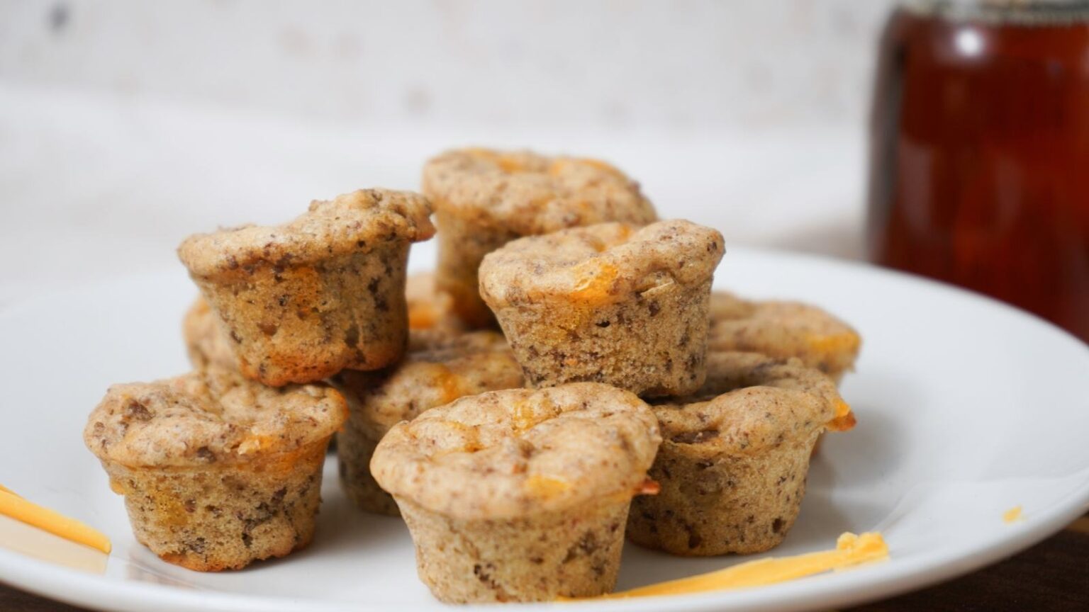 A plate with a stack of small, muffin-shaped baked goods with visible cheese pieces. A jar of liquid and cheese stick are in the background.