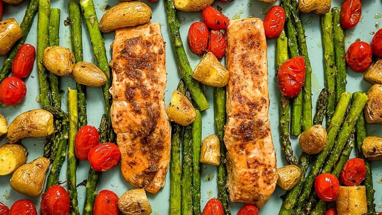 Two baked salmon fillets with roasted asparagus, cherry tomatoes, and halved baby potatoes arranged on a baking sheet.