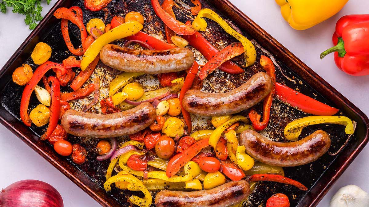 Sheet pan with cooked sausages, cherry tomatoes, sliced red and yellow bell peppers, onions, and garlic cloves.