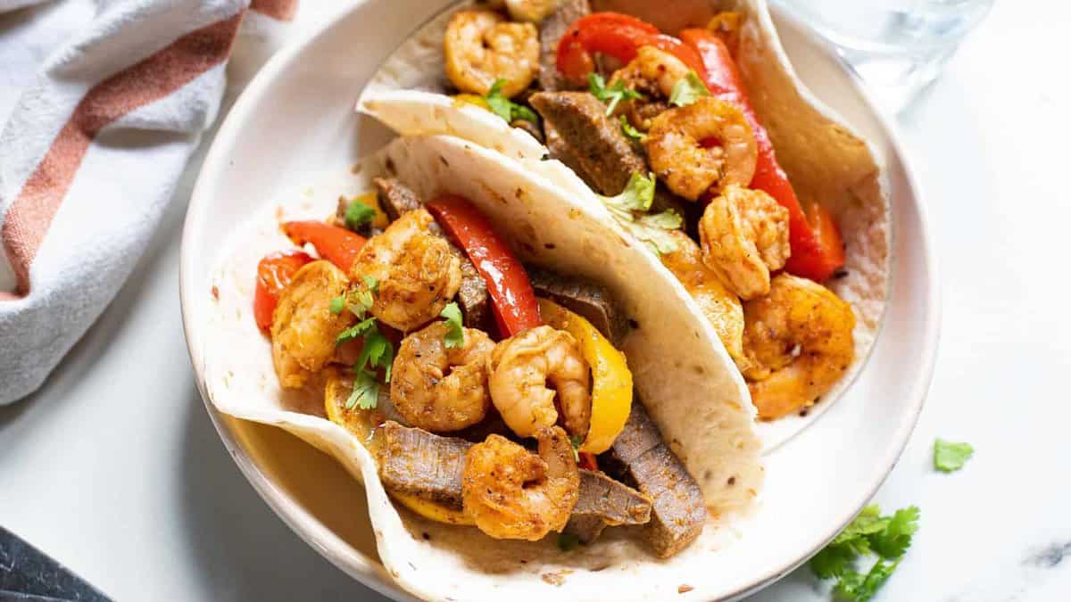 Two flour tortillas filled with shrimp, beef strips, red and yellow bell peppers, and cilantro, served on a round white plate.