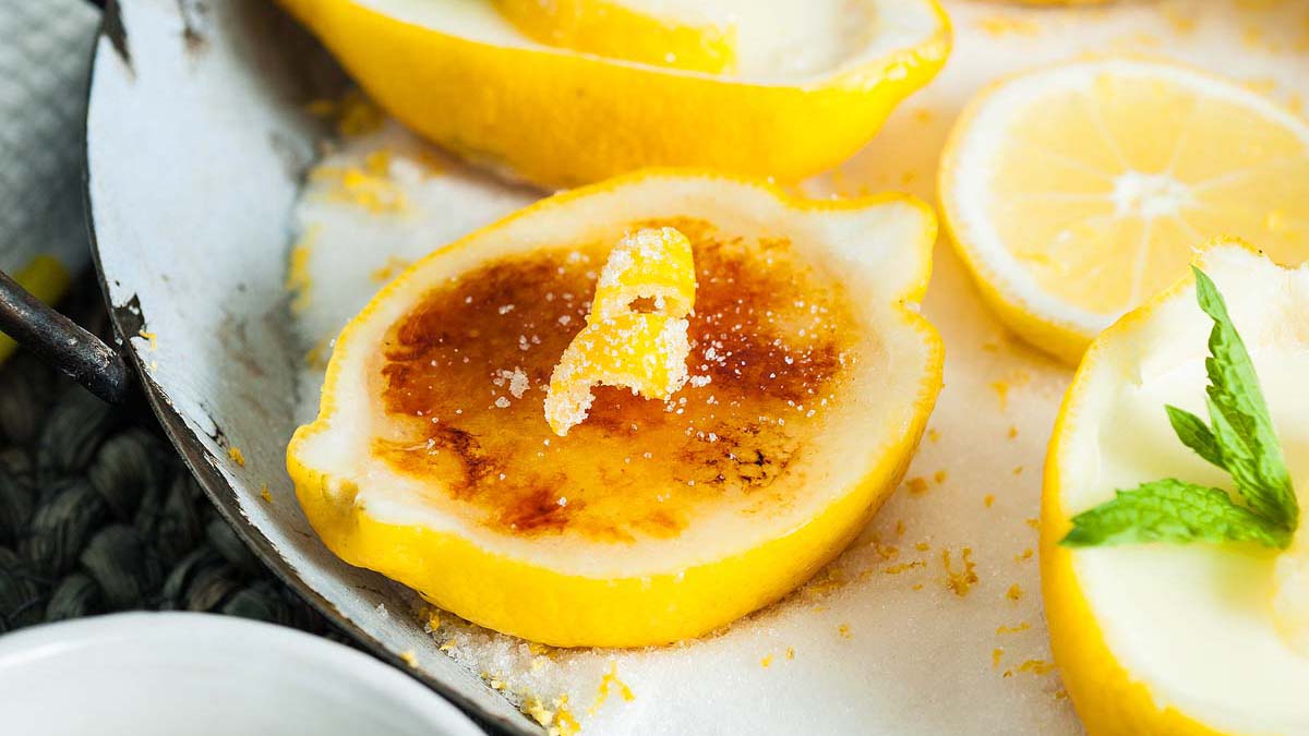 Half a lemon filled with caramelized custard, garnished with a lemon peel twist, sits on a tray with other lemon halves and a mint leaf.