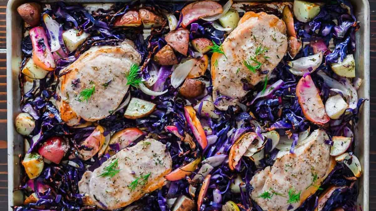 Sheet pan with four baked pork chops, surrounded by roasted purple cabbage, red potatoes, onions, and apple slices, garnished with fresh herbs.