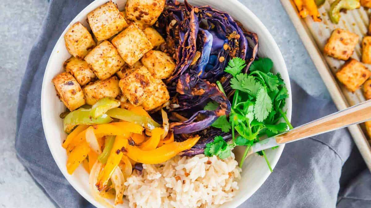 A bowl containing cubed tofu, roasted purple cabbage, sautéed bell peppers, brown rice, and fresh cilantro, with a fork on the side.