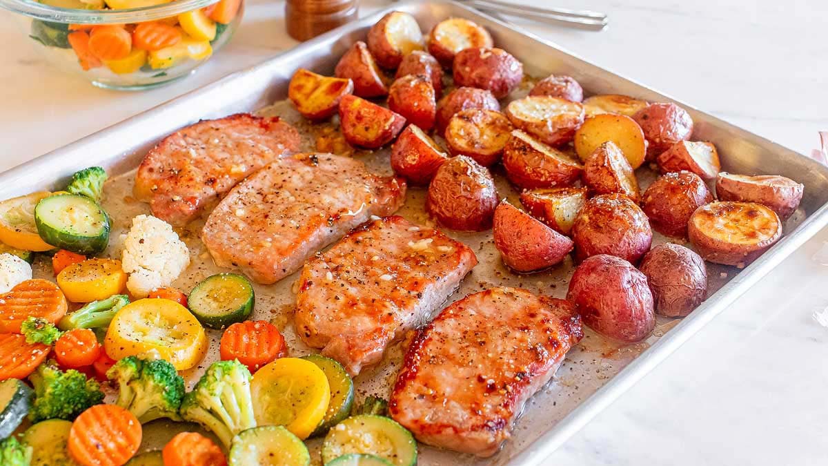 A sheet pan with four cooked pork chops, roasted red potatoes, and mixed vegetables including zucchini, carrots, broccoli, and cauliflower.