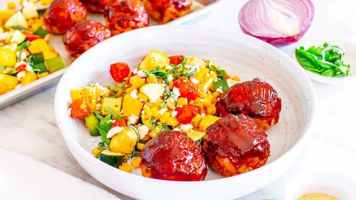 A white bowl with three barbecue-glazed meatballs and a colorful vegetable medley, including corn, tomatoes, zucchini, and cheese. A baking tray and half a red onion are in the background.