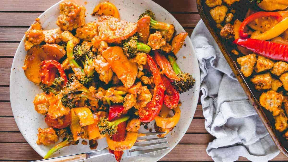 A plate of stir-fried vegetables and chunks of chicken in a sauce, served with a fork on a wooden table, next to a baking sheet with more of the same dish.