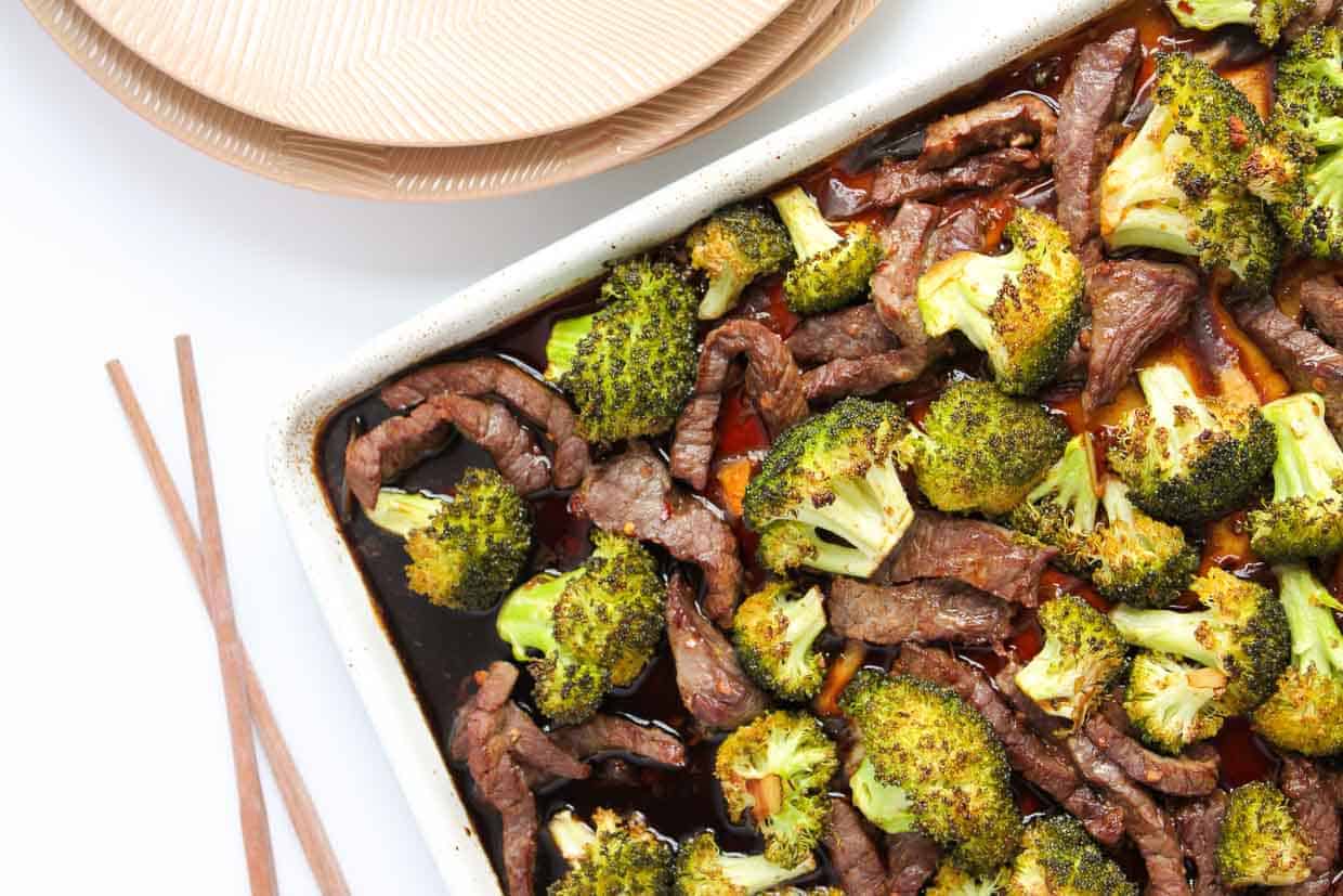 A baking dish filled with cooked beef strips and roasted broccoli next to a stack of plates and a pair of chopsticks.