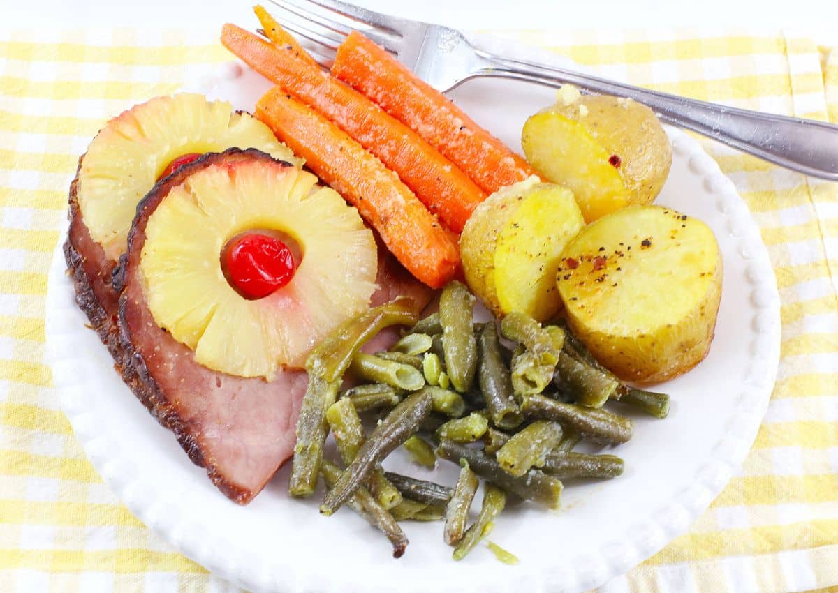 A plate with ham topped with pineapple and a cherry, roasted carrots, potatoes, and green beans, with a fork on a yellow checkered napkin.