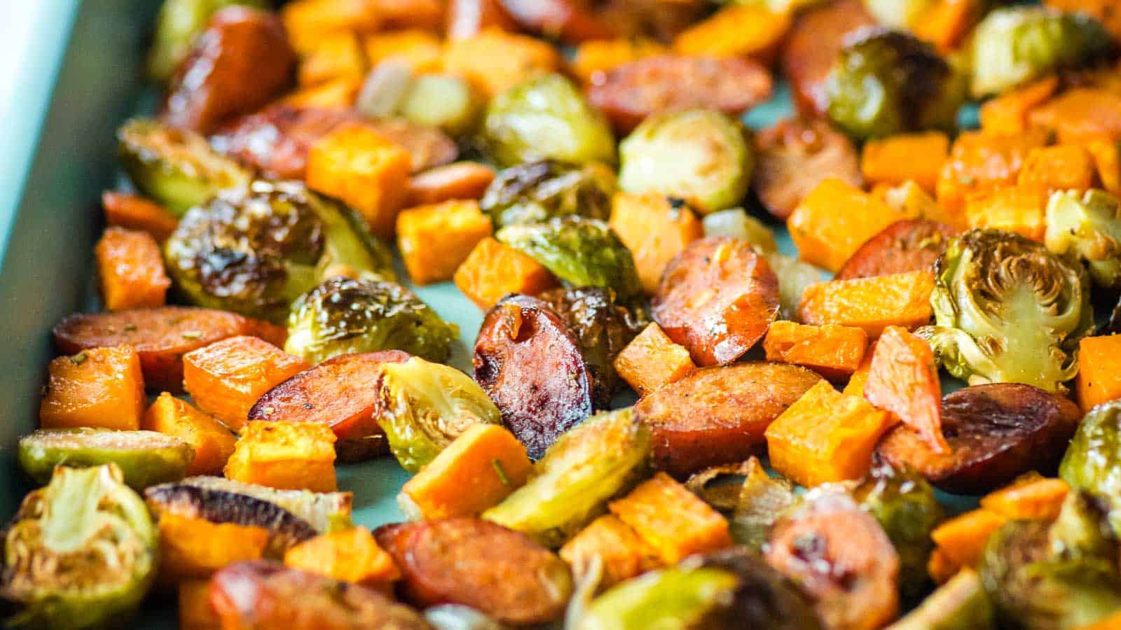 Roasted vegetables and sliced sausage pieces arranged on a baking sheet, including Brussels sprouts and diced sweet potatoes.