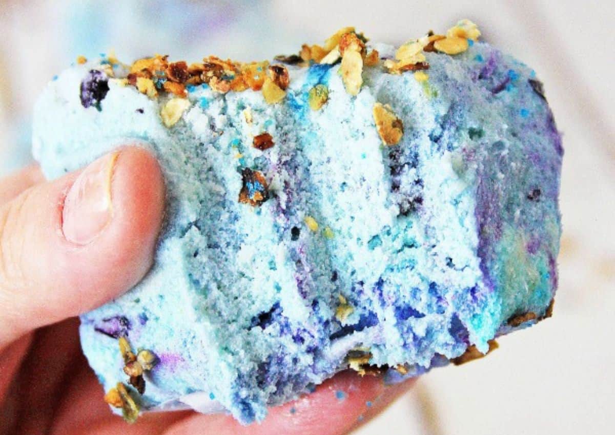 A hand holding a blue and purple bath bomb topped with bits of granola or dried oats.