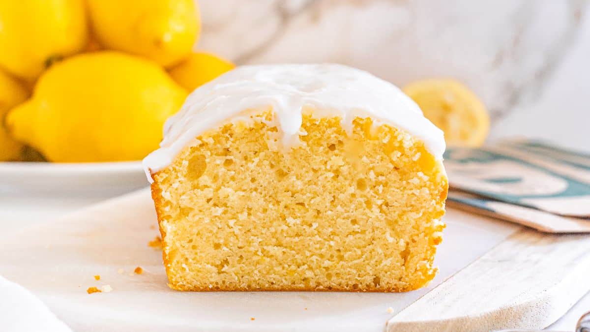 A loaf of lemon drizzle cake with a white icing glaze is sliced in half on a cutting board, with fresh lemons in the background.