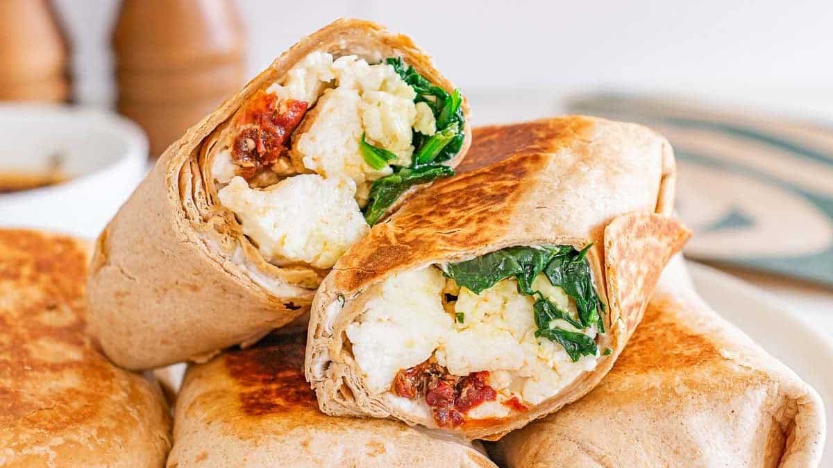 A close-up of toasted breakfast wraps cut in half, showing scrambled eggs, spinach, and sun-dried tomatoes inside a whole wheat tortilla.