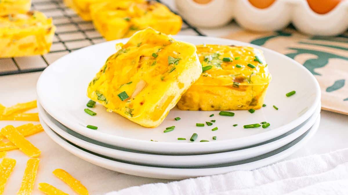 Two square egg muffins garnished with chopped chives are served on stacked white plates, with more egg muffins on a cooling rack in the background.