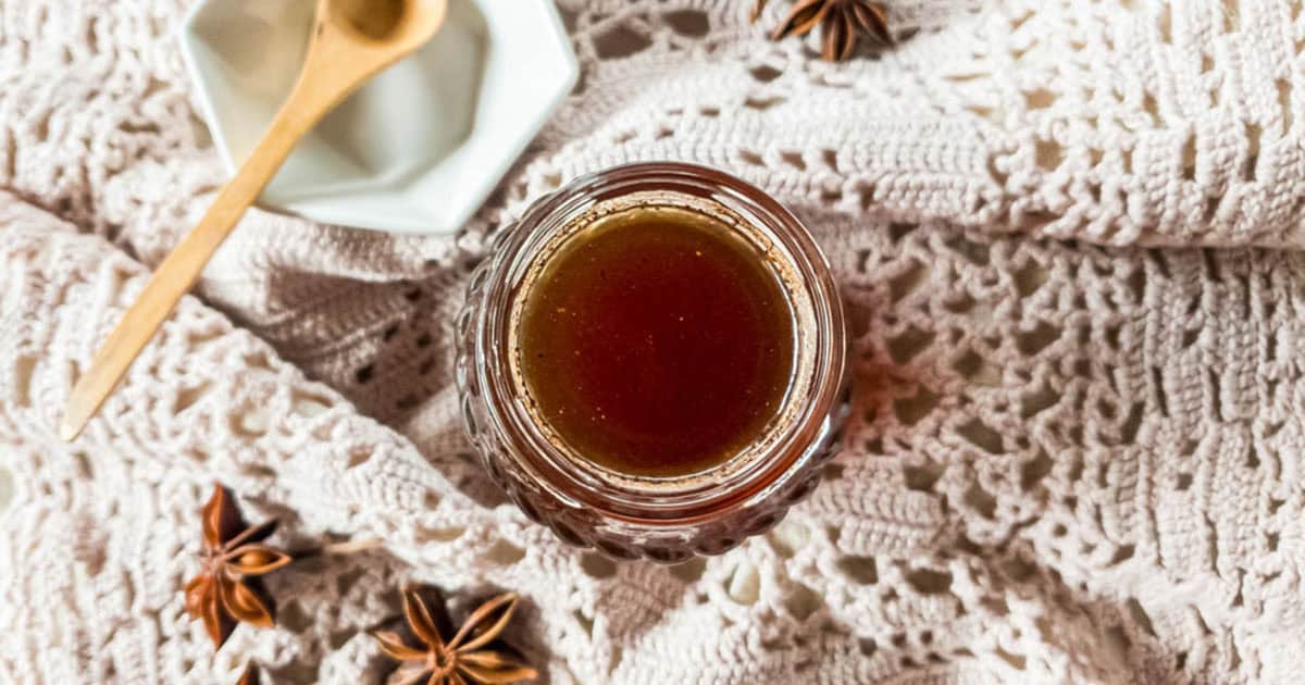 A glass jar filled with dark syrup sits on a crocheted fabric, surrounded by star anise and a small plate with a wooden spoon.