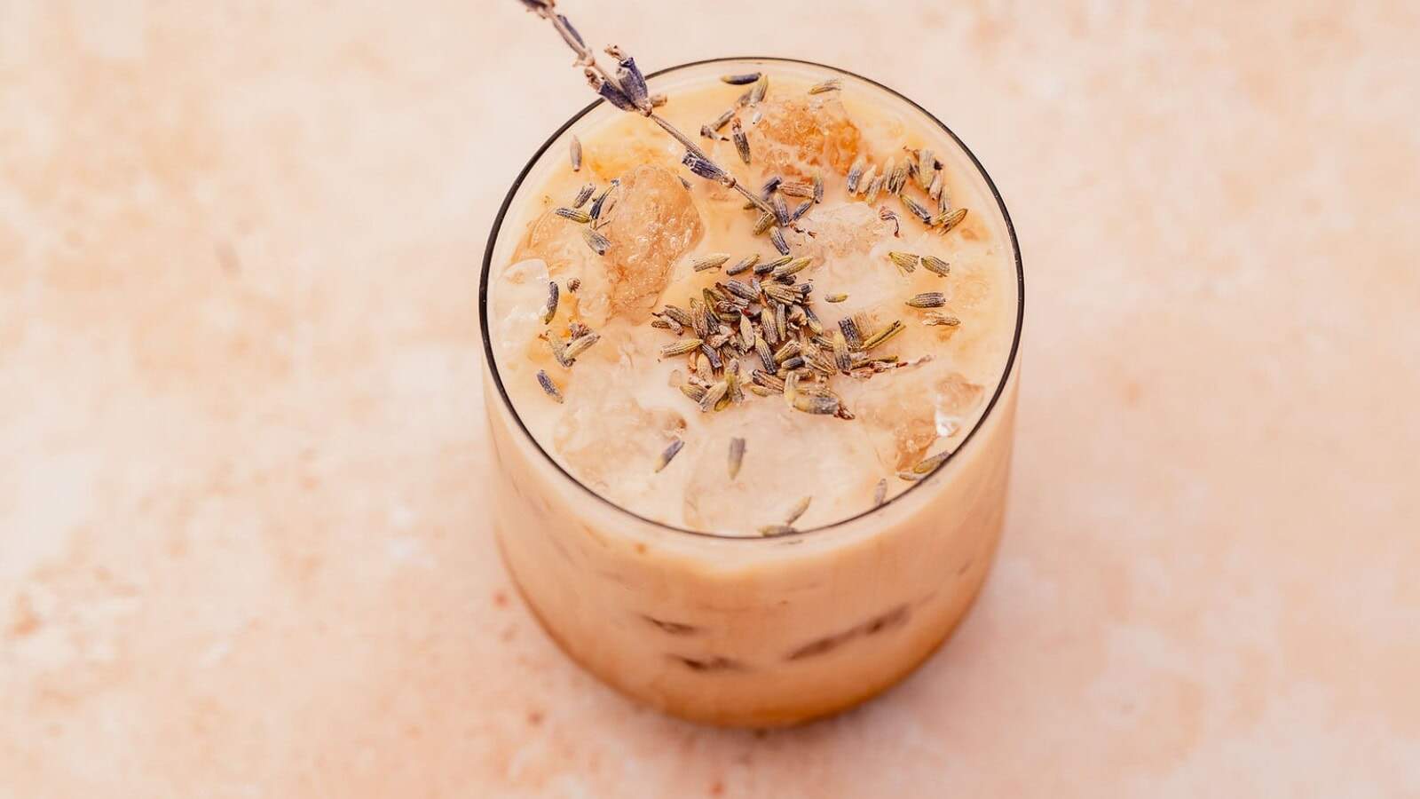 A glass of iced coffee with milk, topped with ice cubes and sprinkled with dried lavender buds, garnished with a lavender sprig.