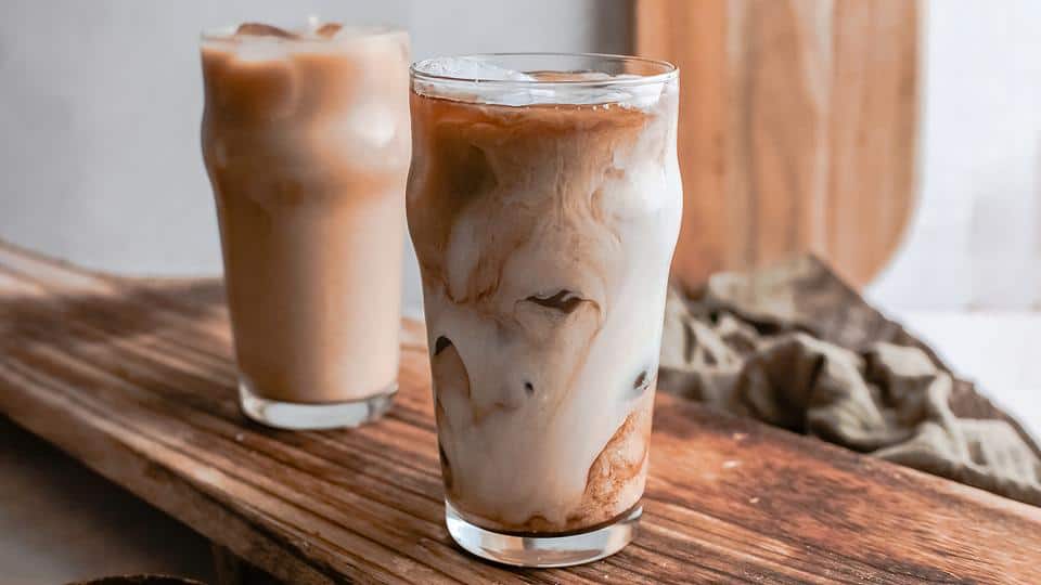 Two glasses of iced coffee with milk sit on a wooden surface; the drink in the foreground shows milk swirling into the coffee.