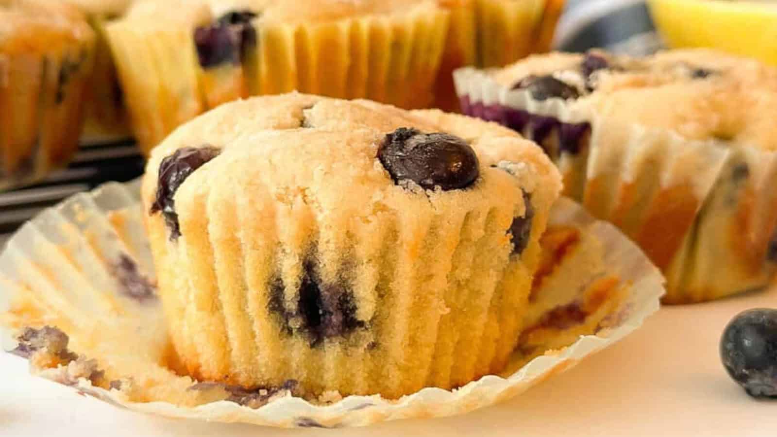 A close-up of a blueberry muffin in a paper liner with visible whole blueberries, surrounded by more muffins in the background.