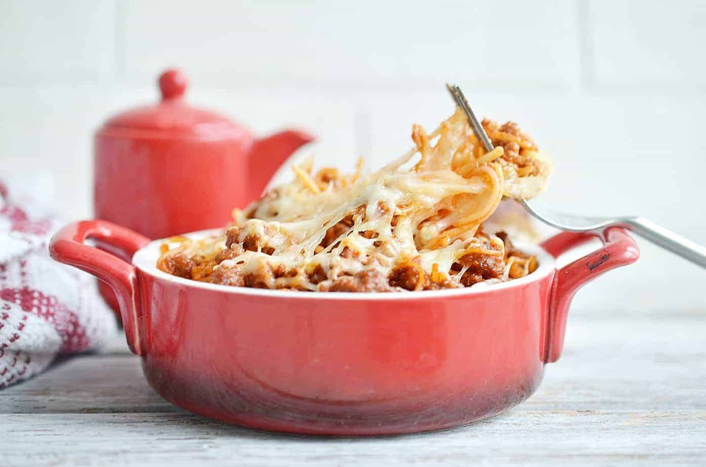 A red ceramic dish filled with cheesy baked pasta and meat sauce, with a fork lifting a serving; a red teapot is in the background.