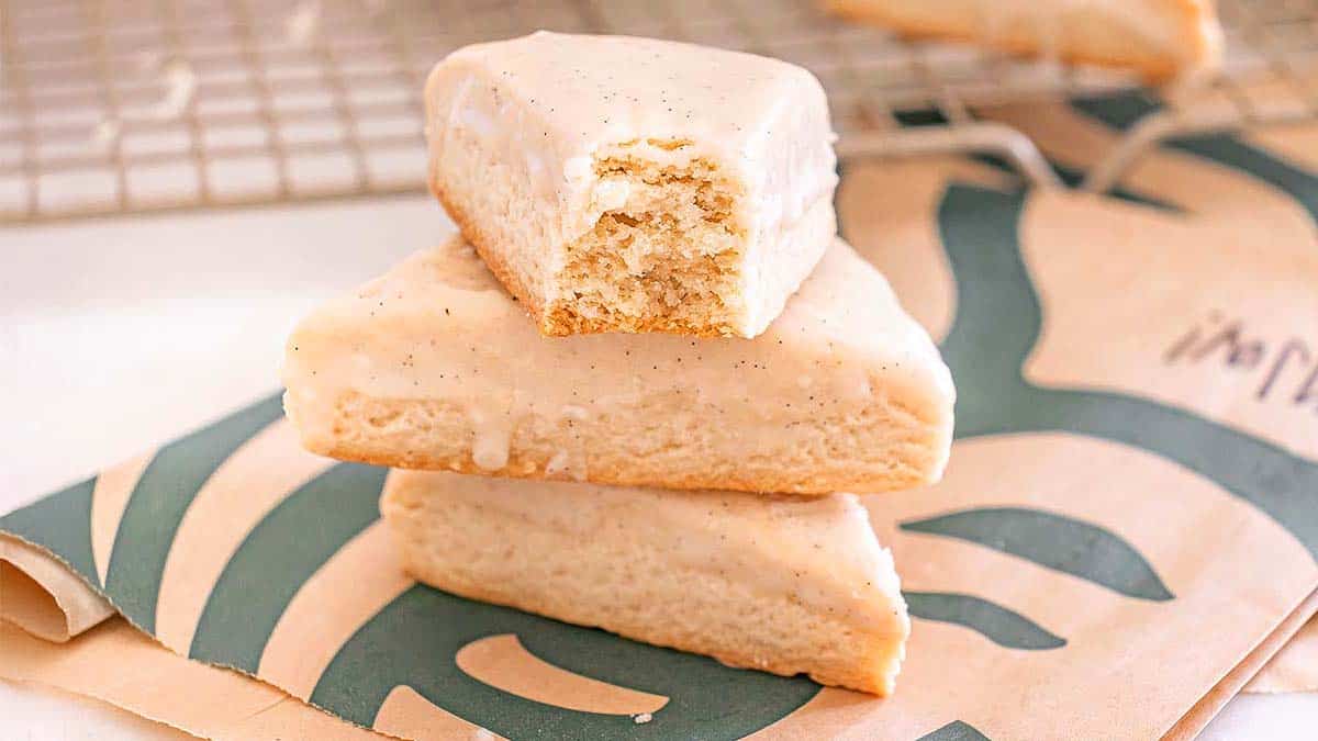 Three vanilla-iced scones are stacked on a branded napkin, with the top scone showing a bite taken out. A cooling rack appears in the background.